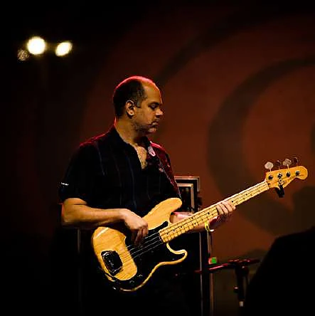 Rodrigo Balduino tocando baixo em um show, iluminado por luz quente de palco